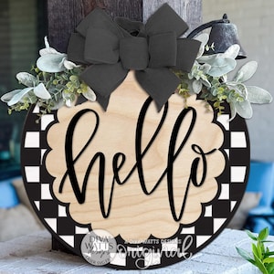 May include: A round wooden door sign featuring a black and white checkered border and a scalloped center with the word hello in black script. The sign is decorated with a large black fabric bow and artificial green foliage at the top.
