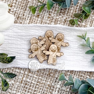 May include: A collection of five wooden figurines depicting Jesus with outstretched arms. The figures are light brown and have etched details. The figures are arranged on a white cloth, surrounded by greenery and a white flower.