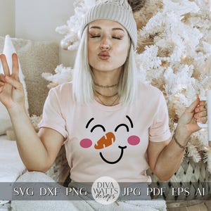 Peut inclure: Une femme portant un t-shirt rose avec un motif de visage de bonhomme de neige souriant. Le bonhomme de neige a des joues roses et un nez de carotte. La femme tient une tasse blanche avec une poign&eacute;e en bois.
