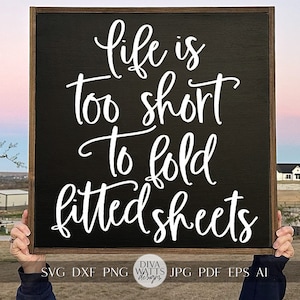 May include: A black wooden sign with white text that reads "Life is too short to fold fitted sheets". The sign is being held up by a person's hands in front of a blurry background of houses and trees.