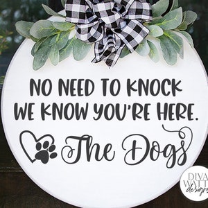 May include: White round wooden sign with black text that reads "No need to knock we know you're here. The Dogs" with a paw print and heart design. The sign is decorated with a black and white checked bow and green leaves.