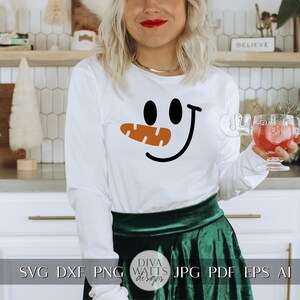 Peut inclure: Un t-shirt blanc &agrave; manches longues avec un motif de visage de bonhomme de neige noir et orange. Le t-shirt est port&eacute; par une personne avec une jupe verte et se trouve dans un d&eacute;cor de cuisine.