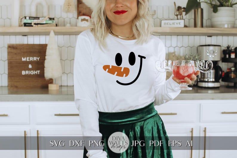 Peut inclure: Un t-shirt blanc &agrave; manches longues avec un motif de visage de bonhomme de neige noir et orange. Le t-shirt est port&eacute; par une personne avec une jupe verte et se trouve dans un d&eacute;cor de cuisine.