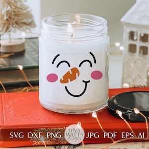Peut inclure: Un bocal de bougie blanc avec un motif de visage de bonhomme de neige. Le bonhomme de neige a des joues roses, un sourire noir et un nez de carotte orange. Le bocal de bougie est pos&eacute; sur un livre rouge avec le texte "SVG DXF PNG JPG PDF EPS AI" imprim&eacute; sur la couverture.
