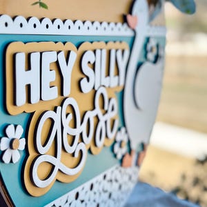 May include: A wooden sign with a light blue background and a yellow border. The sign says "Hey Silly Goat" in white and yellow letters. There are white flowers and a white silhouette of a goat on the sign.