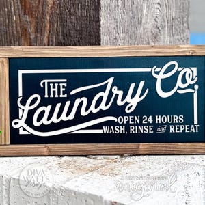 Può includere: Un cartello rettangolare in legno con sfondo blu navy e scritte bianche che recita "The Laundry Co." con "OPEN 24 HOURS WASH, RINSE and REPEAT" sotto. Il cartello è incorniciato in legno marrone chiaro.