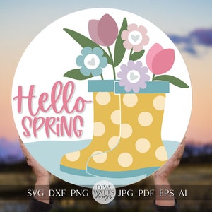 Peut inclure: Une pancarte ronde en bois avec le texte "Hello Spring" en lettres roses. La pancarte représente une paire de bottes de pluie jaunes à pois blancs, tenant un bouquet de fleurs roses, violettes et bleues avec des cœurs blancs au centre de chaque fleur.
