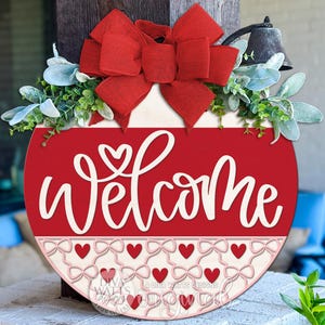 May include: A round, red wooden welcome sign with white script lettering and a heart detail. The sign is decorated with a large red bow, greenery, and a row of hearts and bows. A small bell hangs above the sign.