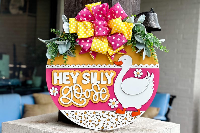 May include: A pink and yellow polka dot bow sits on top of a round wooden door hanger. The door hanger has a white goose with orange feet and the words "Hey Silly Goose" in yellow letters on a pink background.