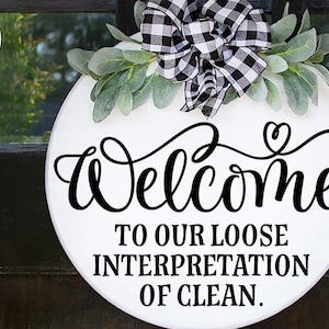 May include: A white round wooden sign with a black and white checkered bow and greenery. The sign says "Welcome to our loose interpretation of clean."