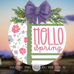 May include: A round wooden sign with a green border and a white background. The sign has a floral pattern on the left side and the text "Hello Spring" in pink on the right side. A purple bow is at the top of the sign.