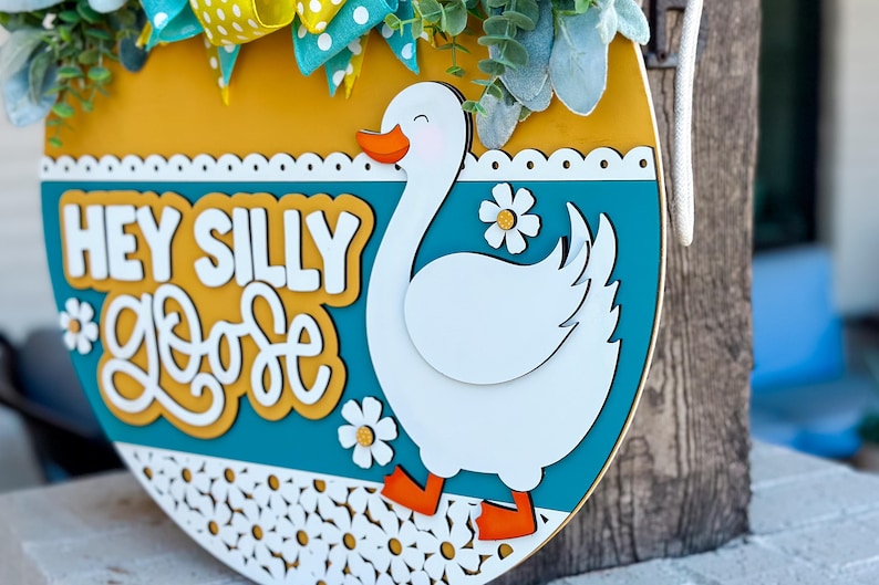 May include: A wooden door hanger with a yellow, teal, and white color scheme. It features a white goose with orange feet and the text "Hey Silly Goose" in white letters on a yellow background.