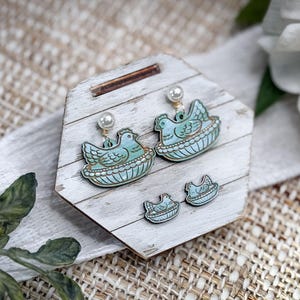 May include: A set of earrings and studs featuring a light blue hen in a nest design. The earrings have a pearl accent. The jewelry is displayed on a white wooden stand with a rustic, farmhouse aesthetic.