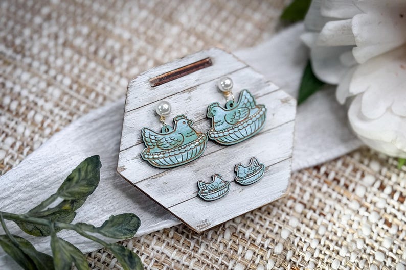 May include: A set of earrings and studs featuring a light blue hen in a nest design. The earrings have a pearl accent. The jewelry is displayed on a white wooden stand with a rustic, farmhouse aesthetic.