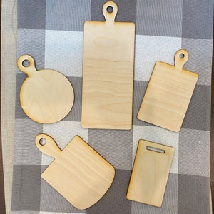 May include: Five unfinished wooden cutting boards of various shapes and sizes. The boards have handles and are arranged on a gray and white checkered fabric.