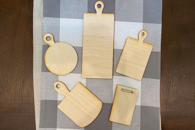 May include: Five unfinished wooden cutting boards of various shapes and sizes. The boards have handles and are arranged on a gray and white checkered fabric.