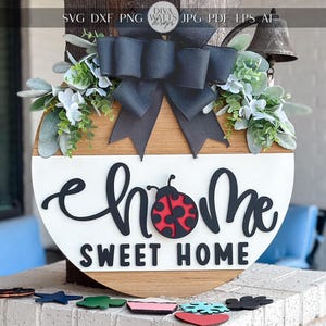 May include: A circular wooden sign with the text "Home Sweet Home" in black script. A red and black ladybird is in the centre. The sign is decorated with a black bow, greenery, and a small bell.