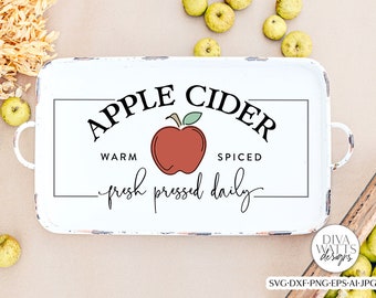 Puede incluir: Una bandeja rectangular blanca con asas, con el texto "APPLE CIDER" sobre una ilustración de una manzana roja. También está presente el texto "WARM SPICED fresh pressed daily". La bandeja está rodeada de manzanas y flores secas.
