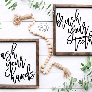 May include: Two wooden signs with black calligraphy on a white background. The signs are on a white wood background with greenery and a wooden bead garland. The text on the signs reads "wash your hands" and "brush your teeth".