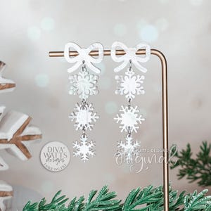 May include: White snowflake earrings with bow details suspended from a gold-coloured display stand. The earrings feature three descending snowflake shapes. The background includes a blurred bokeh effect and decorative snowflakes.