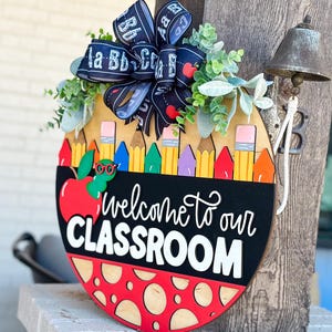 Puede incluir: Un colgador de puerta redondo de madera con una manzana roja, l&aacute;pices de colores y el texto "Bienvenido a nuestra aula". El colgador de puerta est&aacute; decorado con una cinta blanca y negra con las letras "A, B, C, D, E, F" en ella.