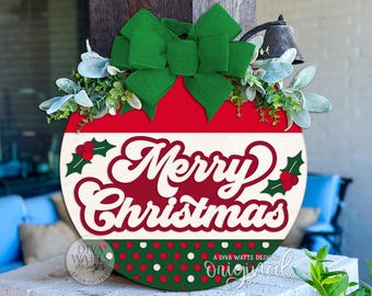 May include: A round Christmas sign with a red and white design, featuring the words "Merry Christmas" in a festive font. It is adorned with a green bow, holly accents, and a small bell. The sign is decorated with a green and red polka dot pattern.
