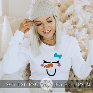 Peut inclure: Un t-shirt blanc &agrave; manches longues avec un motif de visage de bonhomme de neige. Le bonhomme de neige a des joues roses, une bouche noire, un chapeau noir et un n&oelig;ud bleu. Le t-shirt porte le texte "Diva Watts Designs" en bas.