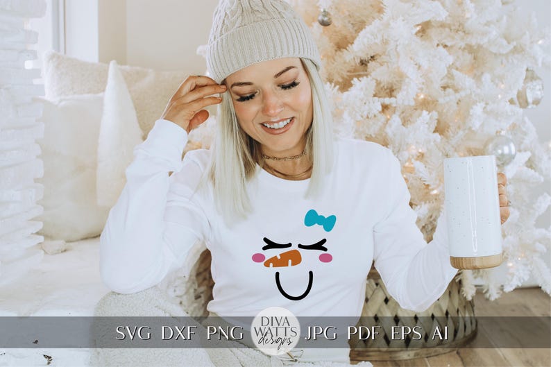 Peut inclure: Un t-shirt blanc &agrave; manches longues avec un motif de visage de bonhomme de neige. Le bonhomme de neige a des joues roses, une bouche noire, un chapeau noir et un n&oelig;ud bleu. Le t-shirt porte le texte "Diva Watts Designs" en bas.