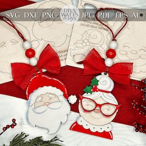 May include: Two unfinished wooden ornaments shaped like Santa Claus and a woman with a white bun. Both ornaments have red bows and are ready to be painted. The ornaments are on a red surface with a green plant in the background.