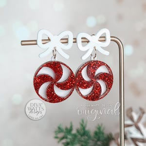 May include: A pair of festive earrings with a red glitter swirl design, and white bow-shaped tops. The earrings are displayed on a gold-coloured stand, ideal for holiday or Christmas themed jewellery.