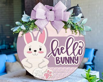 Pode incluir: Uma placa redonda de madeira de Páscoa com uma ilustração de coelho branco e as palavras "hello BUNNY" em roxo. A placa é decorada com um laço lavanda e vegetação, perfeita para decoração de primavera.