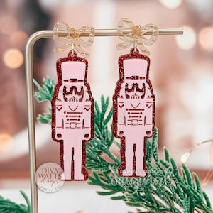 Könnte beinhalten: Rosa und rot glitzernde Nussknacker-Ohrringe mit Schleifenakzenten. Die Ohrringe sind lasergeschnitten und haben ein festliches Design. Die Ohrringe werden auf einem silbernen Ständer mit Grün im Hintergrund präsentiert. Ein Accessoire zum Thema Feiertage.