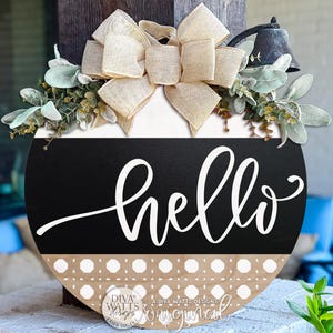 Puede incluir: Letrero redondo de madera con la palabra "hello" en escritura blanca. El letrero tiene un centro negro, una parte superior blanca y una base estampada. Está adornado con un lazo beige y vegetación. Una pequeña campana está adjunta.