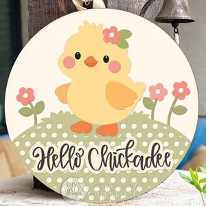 May include: Round wooden sign featuring a cartoon chick with a flower crown, standing on a dotted green hill with pink flowers. The sign reads "Hello Chickadee" in a script font. Decorative Easter-themed item.