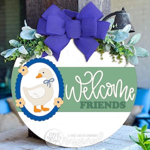 May include: A circular welcome sign with a blue bow and foliage. The sign features a white goose in a blue oval frame with orange flowers. The words "Welcome Friends" are written in white script across the sign.