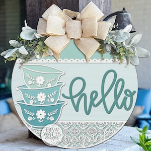 May include: A round wooden welcome sign with a light blue background and the word "hello" in teal script. The sign features stacked teal bowls with floral designs and is decorated with a beige bow and greenery.