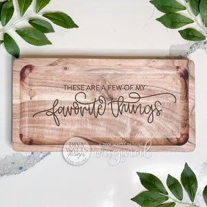 May include: A rectangular wooden serving tray with the phrase "THESE ARE A FEW OF MY favorite things" etched into the wood. The tray has a light brown color and a recessed area. Green leaves are in the background.