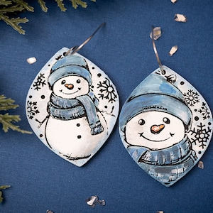 May include: A pair of blue and white snowman earrings with snowflake details. The earrings are made of polymer clay and have a silver hook.