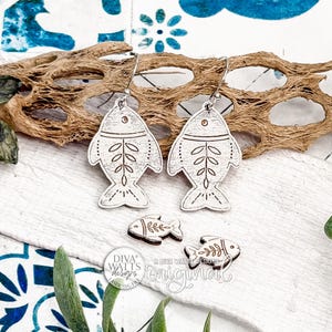 May include: A pair of white fish-shaped earrings with etched leaf and dot details, displayed with two smaller matching fish and a piece of driftwood. The earrings are on a white surface with green foliage and blue patterned fabric.