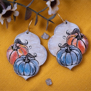 May include: A pair of pumpkin-themed earrings with a white background and a distressed look. The earrings feature two pumpkins, one orange and one blue, with a black outline. The pumpkins have a watercolor effect and are painted in a realistic style.