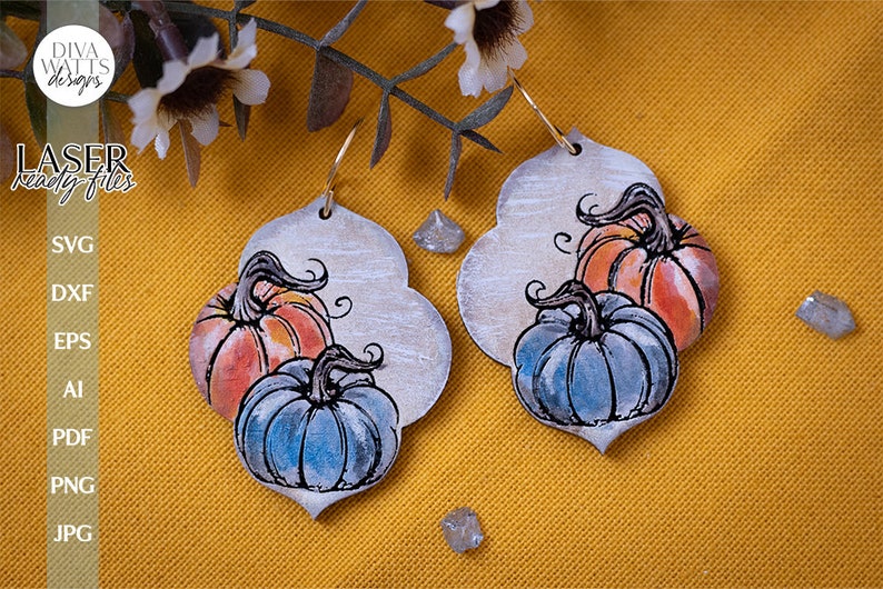 May include: A pair of pumpkin-themed earrings with a white background and a distressed look. The earrings feature two pumpkins, one orange and one blue, with a black outline. The pumpkins have a watercolor effect and are painted in a realistic style.