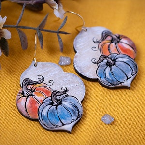 May include: A pair of silver-toned earrings with a pumpkin design. The pumpkins are painted in shades of orange, blue, and green. The earrings are hanging from gold-toned hooks.