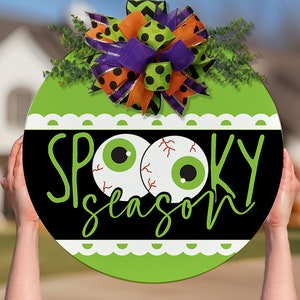 May include: A green and black round door hanger with a white scalloped edge. The door hanger has two googly eyes and the text "Spooky Season" in black script.