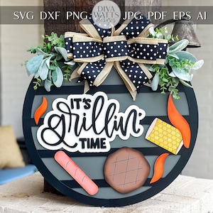 May include: A round, black and gray wooden sign with the text "It's Grillin' Time" in white script. The sign features illustrations of a sausage, steak, corn, and flames. A black and white polka dot bow and greenery adorn the top.