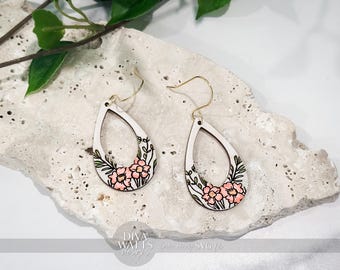 May include: Teardrop-shaped earrings with a floral design. The earrings are white with a black outline and feature pink flowers and green leaves. The earring hooks are gold-colored.