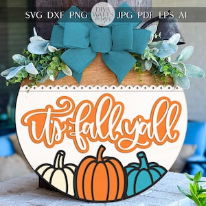 May include: A round wooden sign with the text "It's fall y'all" in orange and white. The sign features three pumpkins in white, orange, and teal. A teal bow and greenery adorn the top.