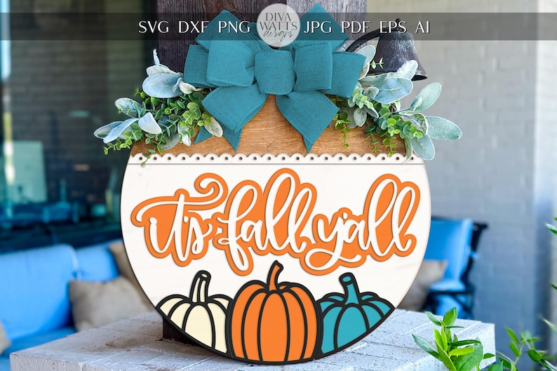 May include: A round wooden sign with the text "It's fall y'all" in orange and white. The sign features three pumpkins in white, orange, and teal. A teal bow and greenery adorn the top.