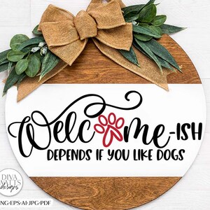 Pode incluir: Uma placa redonda de madeira com um laço de juta marrom e folhas verdes. A placa diz "Welcome-ish Depends If You Like Dogs" em texto preto com uma pegada vermelha.