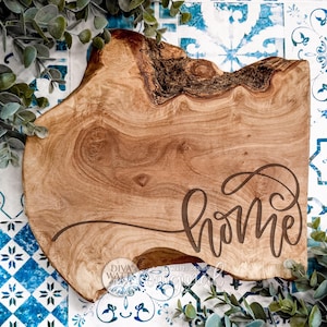 May include: A rustic wooden serving board with a natural edge. The board is light brown with a dark brown inscription of the word "home" in a cursive font. Greenery and a blue and white patterned surface surround the board.