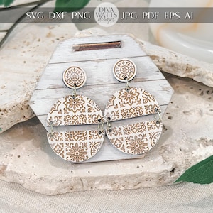 Könnte beinhalten: Holzohrringe mit floralem Muster. Die Ohrringe haben ein kreisförmiges Design mit einem detaillierten, lasergeschnittenen Blumenmotiv in hellbrauner Farbe. Die Ohrringe hängen an einem kleinen, runden Stecker. Sie werden auf einer weißen Holzoberfläche präsentiert.